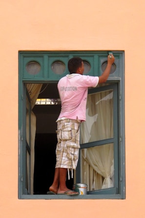 A person wearing a pink shirt and plaid shorts is balancing on the edge of an open window while painting the upper part of the frame. The window has a green frame with multiple small circular panes at the top. There is a paint can and brush being used for painting. The curtains inside the room are drawn, and the overall wall color is a peach or light orange.