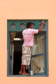 A person wearing a pink shirt and plaid shorts is balancing on the edge of an open window while painting the upper part of the frame. The window has a green frame with multiple small circular panes at the top. There is a paint can and brush being used for painting. The curtains inside the room are drawn, and the overall wall color is a peach or light orange.