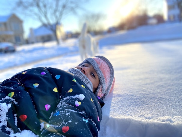A child is bundled up in a colorful blanket featuring heart patterns, lying on a snow-covered surface. The child wears a knit hat, and the background shows a snowy landscape with blurred houses and a tree. The sunlight casts a warm glow, creating a bright winter scene.