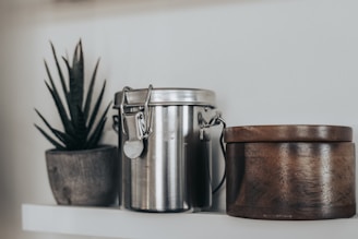 A wooden container, a metal canister with a clamp lid, and a small potted plant are arranged on a white shelf. The plant is in a rustic pot, and the canister reflects light with its shiny surface.