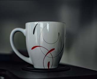 A ceramic mug with a minimalist design featuring red and black swirls. The handle is visible to the left, and the mug is placed on a dark surface with a blurred gray background.
