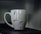 A ceramic mug with a minimalist design featuring red and black swirls. The handle is visible to the left, and the mug is placed on a dark surface with a blurred gray background.