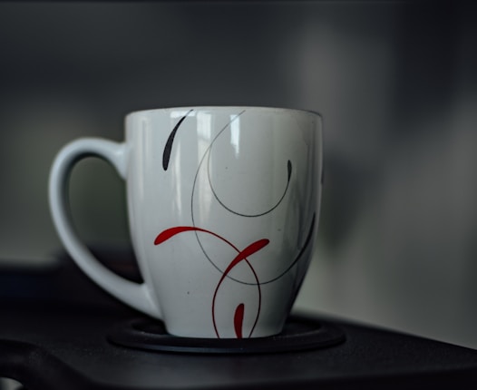 A ceramic mug with a minimalist design featuring red and black swirls. The handle is visible to the left, and the mug is placed on a dark surface with a blurred gray background.