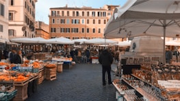A bustling outdoor market scene with various stalls selling fruits, vegetables, and other goods under large white canopies. People are shopping and walking along the cobblestone street lined with crates of produce. In the background, a row of historic European buildings provides a picturesque urban setting.