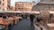 A bustling outdoor market scene with various stalls selling fruits, vegetables, and other goods under large white canopies. People are shopping and walking along the cobblestone street lined with crates of produce. In the background, a row of historic European buildings provides a picturesque urban setting.