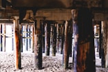 Wooden support beams under a pier are visible, providing a view of the sandy ground beneath. The beams vary in color and texture, with some showing signs of wear and watermarks.