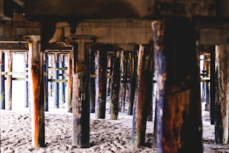 Wooden support beams under a pier are visible, providing a view of the sandy ground beneath. The beams vary in color and texture, with some showing signs of wear and watermarks.