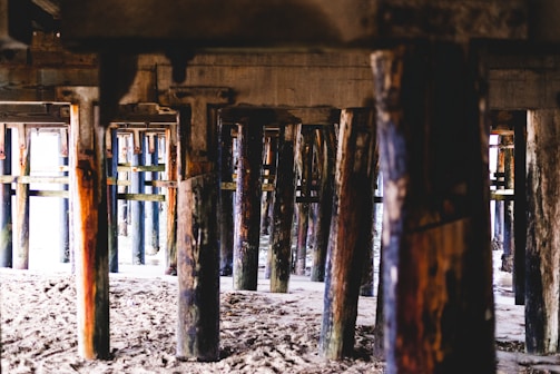 Wooden support beams under a pier are visible, providing a view of the sandy ground beneath. The beams vary in color and texture, with some showing signs of wear and watermarks.