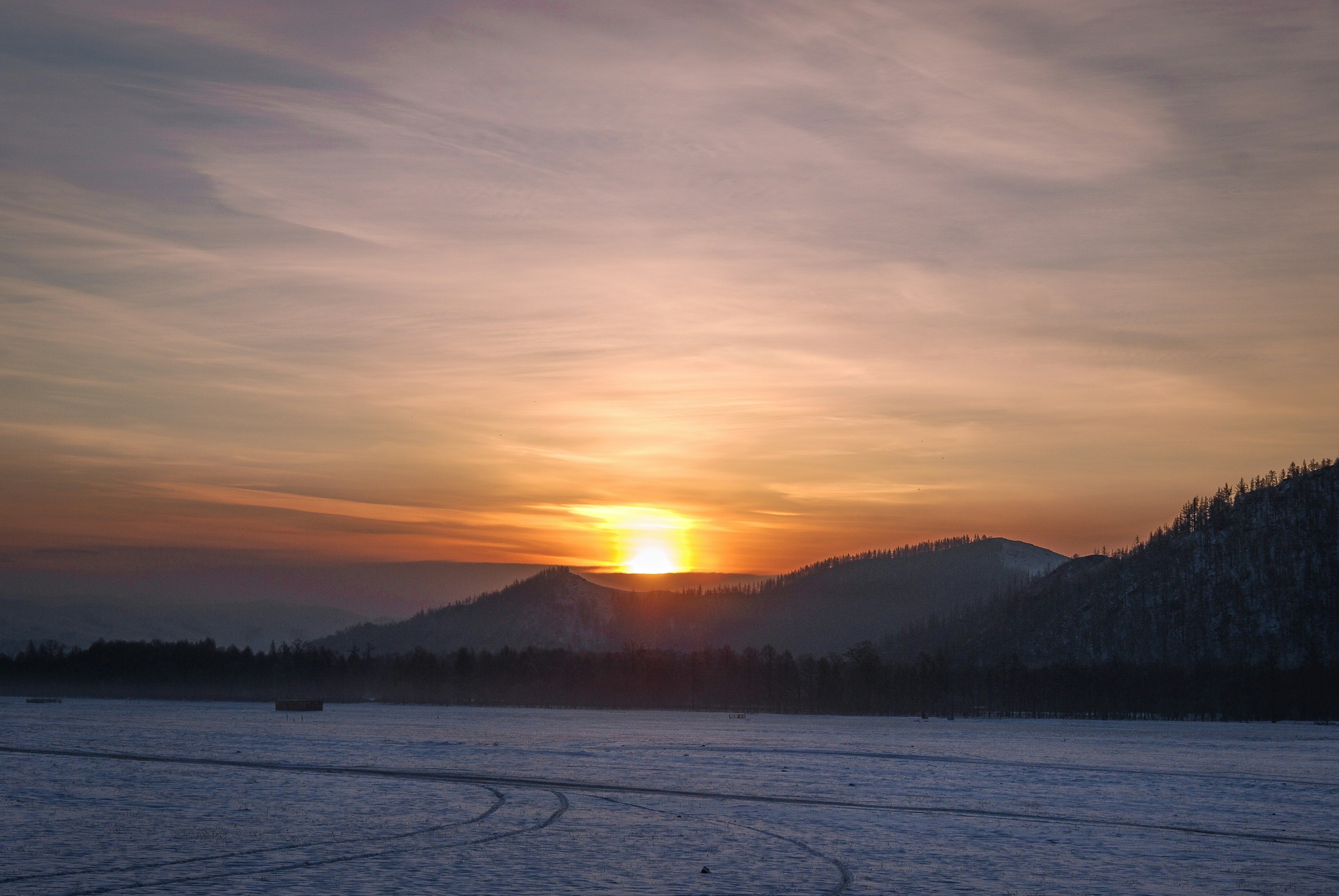 body of water near mountain during sunset, This is a winter sunrise in Mongolia, it is the sun rising in between forest, mountains, clouds, and the flat endless landscapes. 