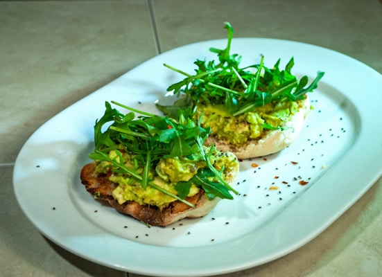 A white plate holds two slices of toasted bread topped with a generous spread of creamy avocado and garnished with fresh arugula leaves. The arrangement is sprinkled with black and red pepper flakes, adding a touch of spice and color to the presentation. The background showcases a simple tiled surface.