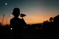 A person with a cap is holding a camera or camcorder, capturing the scene as the sun sets in the background. The surroundings are dimly lit with street lights and what appears to be an industrial structure in the distance.
