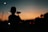 A person with a cap is holding a camera or camcorder, capturing the scene as the sun sets in the background. The surroundings are dimly lit with street lights and what appears to be an industrial structure in the distance.