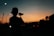 A person with a cap is holding a camera or camcorder, capturing the scene as the sun sets in the background. The surroundings are dimly lit with street lights and what appears to be an industrial structure in the distance.