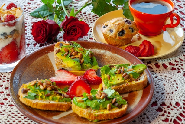 A beautifully arranged breakfast featuring avocado toast topped with pine nuts and cilantro, accompanied by sliced strawberries. On the side, a glass parfait with layers of yogurt, granola, and raspberries is visible. A blueberry muffin sits on a small plate beside a bright red coffee cup filled with black coffee. Two red roses are placed in the background on a lace tablecloth.