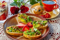 A beautifully arranged breakfast featuring avocado toast topped with pine nuts and cilantro, accompanied by sliced strawberries. On the side, a glass parfait with layers of yogurt, granola, and raspberries is visible. A blueberry muffin sits on a small plate beside a bright red coffee cup filled with black coffee. Two red roses are placed in the background on a lace tablecloth.