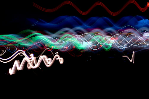 Vibrant, wavy light streaks in a variety of colors including green, blue, red, and white move dynamically against a dark background, creating an abstract and energetic pattern.