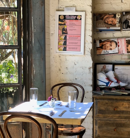A cozy corner in a café features a small table set for two with wooden chairs. A menu poster about choking hangs on a white brick wall. Nearby, shelves hold magazines and a pair of vintage roller skates. A window to the left shows greenery outside, allowing natural light to flood the space.