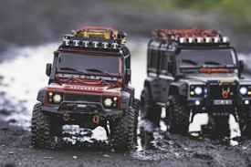 Two toy off-road vehicles are positioned on a muddy path. The vehicle in the foreground is red with detailed features including headlights and a winch, while the one slightly blurred in the background is black. Both have large tires and roof racks loaded with gear, suggesting readiness for a rugged adventure.