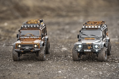 Two detailed scale model Land Rover Defender SUVs are positioned on a rugged, gravelly terrain. One is brown with black accents, while the other is silver with similar black detailing. Both models have roof racks equipped with supplies and floodlights, showcasing intricate craftsmanship. The scene evokes a sense of adventure and off-road exploration.