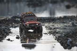 A toy SUV with rugged tires and a roof rack, set in a shallow, reflective puddle surrounded by rocky and muddy terrain. The vehicle appears detailed, with headlights and a grill visible, resembling an adventure or off-road model.