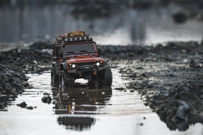 A toy SUV with rugged tires and a roof rack, set in a shallow, reflective puddle surrounded by rocky and muddy terrain. The vehicle appears detailed, with headlights and a grill visible, resembling an adventure or off-road model.