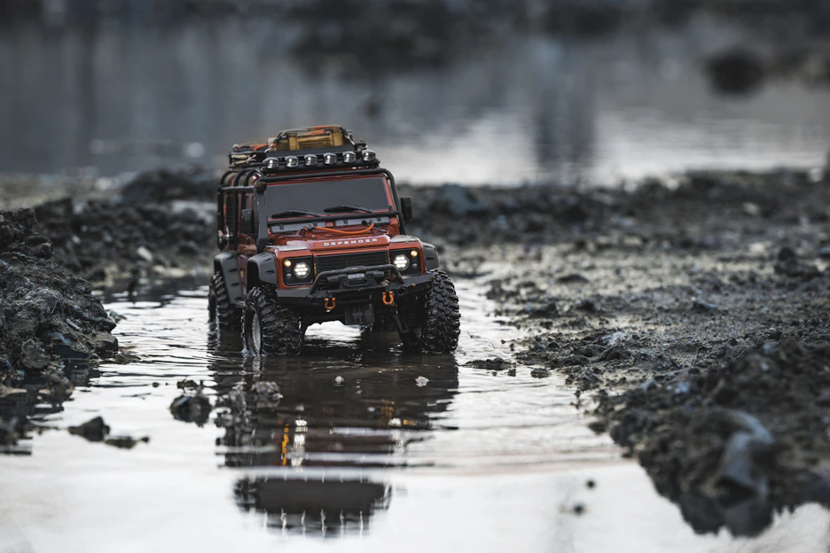 A rugged RC crawler climbing over rocky terrain with a detailed die cast car model beside it.
