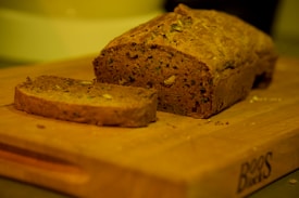 A loaf of brown bread and a sliced piece are on a wooden cutting board. The bread appears to contain nuts and seeds, adding texture and variety to its appearance. The image is warmly lit, creating a cozy and inviting atmosphere.