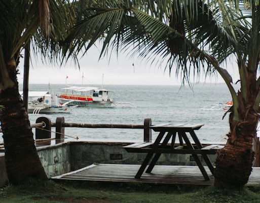 A wooden picnic table is positioned on a patio overlooking the sea. Two palm trees frame the view of the ocean where boats are docked, with one prominently displaying the name Cebu. The sky is overcast, creating a moody and tranquil atmosphere.