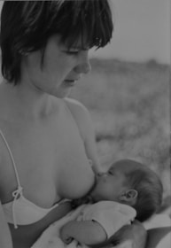 A black and white photograph of a woman wearing a bikini top and looking down at a baby who is breastfeeding. The baby is cradled in the woman's arms, creating a tender and intimate scene.