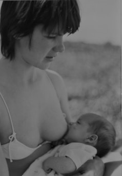 A black and white photograph of a woman wearing a bikini top and looking down at a baby who is breastfeeding. The baby is cradled in the woman's arms, creating a tender and intimate scene.