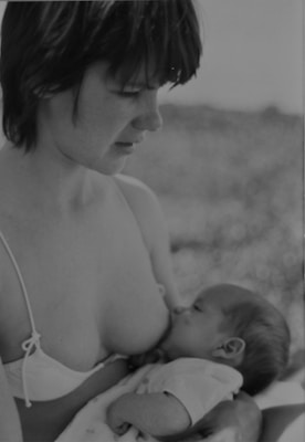 A black and white photograph of a woman wearing a bikini top and looking down at a baby who is breastfeeding. The baby is cradled in the woman's arms, creating a tender and intimate scene.