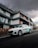 A sleek white electric car is parked on a road in front of a modern, multi-story building. The building features a minimalist architectural design with clean lines and a combination of dark and wooden panels. The sky is overcast, creating a dramatic backdrop with thick, dark clouds.