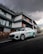 A sleek white electric car is parked on a road in front of a modern, multi-story building. The building features a minimalist architectural design with clean lines and a combination of dark and wooden panels. The sky is overcast, creating a dramatic backdrop with thick, dark clouds.