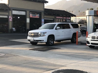 A white Chevrolet Suburban is parked at a gas station. The vehicle is near the fuel pumps, with a garage or service center visible in the background. Another white car is partially visible on the right. Palm trees and mountains are faintly seen in the distance under a soft sky.