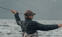 A person wearing a camouflage cap and a black puffy jacket is holding a fishing rod, actively casting a line into a large body of water. The background is composed of blurred mountains and a cloudy sky, conveying an outdoor, nature setting.