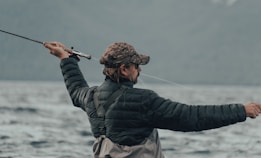 A person wearing a camouflage cap and a black puffy jacket is holding a fishing rod, actively casting a line into a large body of water. The background is composed of blurred mountains and a cloudy sky, conveying an outdoor, nature setting.