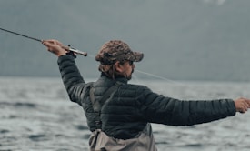 A person wearing a camouflage cap and a black puffy jacket is holding a fishing rod, actively casting a line into a large body of water. The background is composed of blurred mountains and a cloudy sky, conveying an outdoor, nature setting.