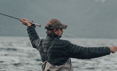 A person wearing a camouflage cap and a black puffy jacket is holding a fishing rod, actively casting a line into a large body of water. The background is composed of blurred mountains and a cloudy sky, conveying an outdoor, nature setting.