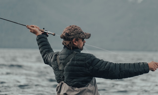 A person wearing a camouflage cap and a black puffy jacket is holding a fishing rod, actively casting a line into a large body of water. The background is composed of blurred mountains and a cloudy sky, conveying an outdoor, nature setting.