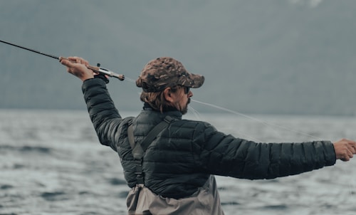A person wearing a camouflage cap and a black puffy jacket is holding a fishing rod, actively casting a line into a large body of water. The background is composed of blurred mountains and a cloudy sky, conveying an outdoor, nature setting.
