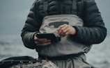 A person wearing a dark padded jacket and a light gray waterproof apron stands by a body of water. They are holding a small black device with both hands, appearing to be focused on it. The background is blurred, showing a calm and cloudy waterscape.