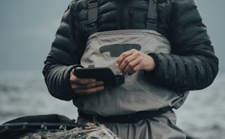 A person wearing a dark padded jacket and a light gray waterproof apron stands by a body of water. They are holding a small black device with both hands, appearing to be focused on it. The background is blurred, showing a calm and cloudy waterscape.