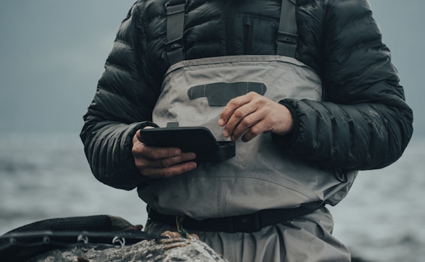 A person wearing a dark padded jacket and a light gray waterproof apron stands by a body of water. They are holding a small black device with both hands, appearing to be focused on it. The background is blurred, showing a calm and cloudy waterscape.