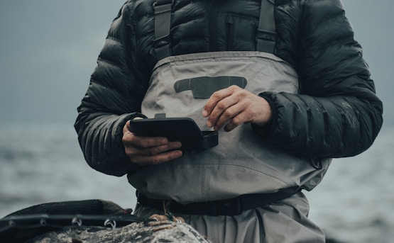 A person wearing a dark padded jacket and a light gray waterproof apron stands by a body of water. They are holding a small black device with both hands, appearing to be focused on it. The background is blurred, showing a calm and cloudy waterscape.