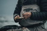 A person wearing a dark puffy jacket and grey overalls is holding a small box, possibly containing fishing tackle. The scene suggests a focus on preparation or maintenance, with hands showing signs of dirt or wear. The background is blurred, likely depicting a natural environment such as a river or lake.