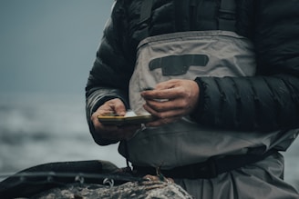 A person wearing a dark puffy jacket and grey overalls is holding a small box, possibly containing fishing tackle. The scene suggests a focus on preparation or maintenance, with hands showing signs of dirt or wear. The background is blurred, likely depicting a natural environment such as a river or lake.