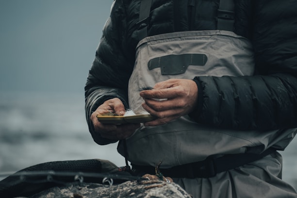 A person wearing a dark puffy jacket and grey overalls is holding a small box, possibly containing fishing tackle. The scene suggests a focus on preparation or maintenance, with hands showing signs of dirt or wear. The background is blurred, likely depicting a natural environment such as a river or lake.