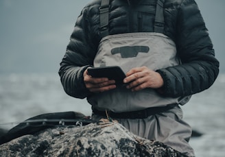 A person is wearing waterproof fishing gear and a padded jacket, standing on a rocky surface near a body of water, holding and looking at a smartphone or similar device.