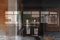 A complex, layered image combining elements of both exterior and interior architectural spaces. The scene includes visible brick walls, glass windows with reflections, and a mix of natural and artificial lighting. Various materials and textures create depth, while furniture and building debris suggest disuse or abandonment.