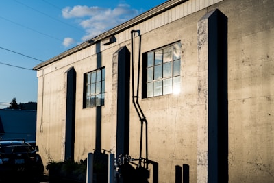 A sunlit industrial building with textured walls and two large rectangular windows casting shadows. The sunlight reflects off the windows creating a bright glare. A car is partially visible in front of the building. The scene includes power lines and a blue sky with scattered clouds.